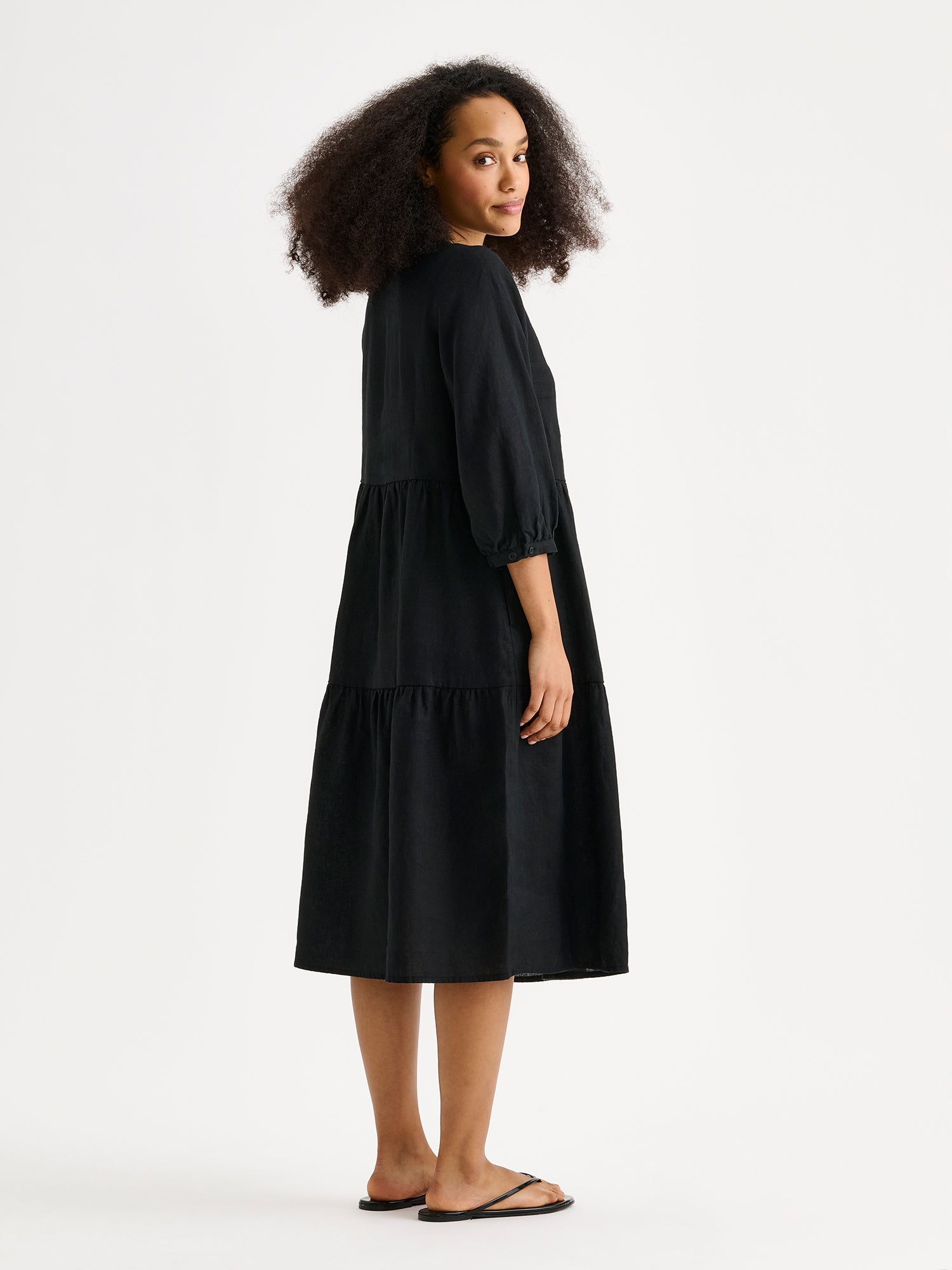 Nanso women's SARALIVIA linen dress in black features charming layered ruffles, 3/4 sleeves, side pockets and a V-neckline. Pictured on a model from the backside.