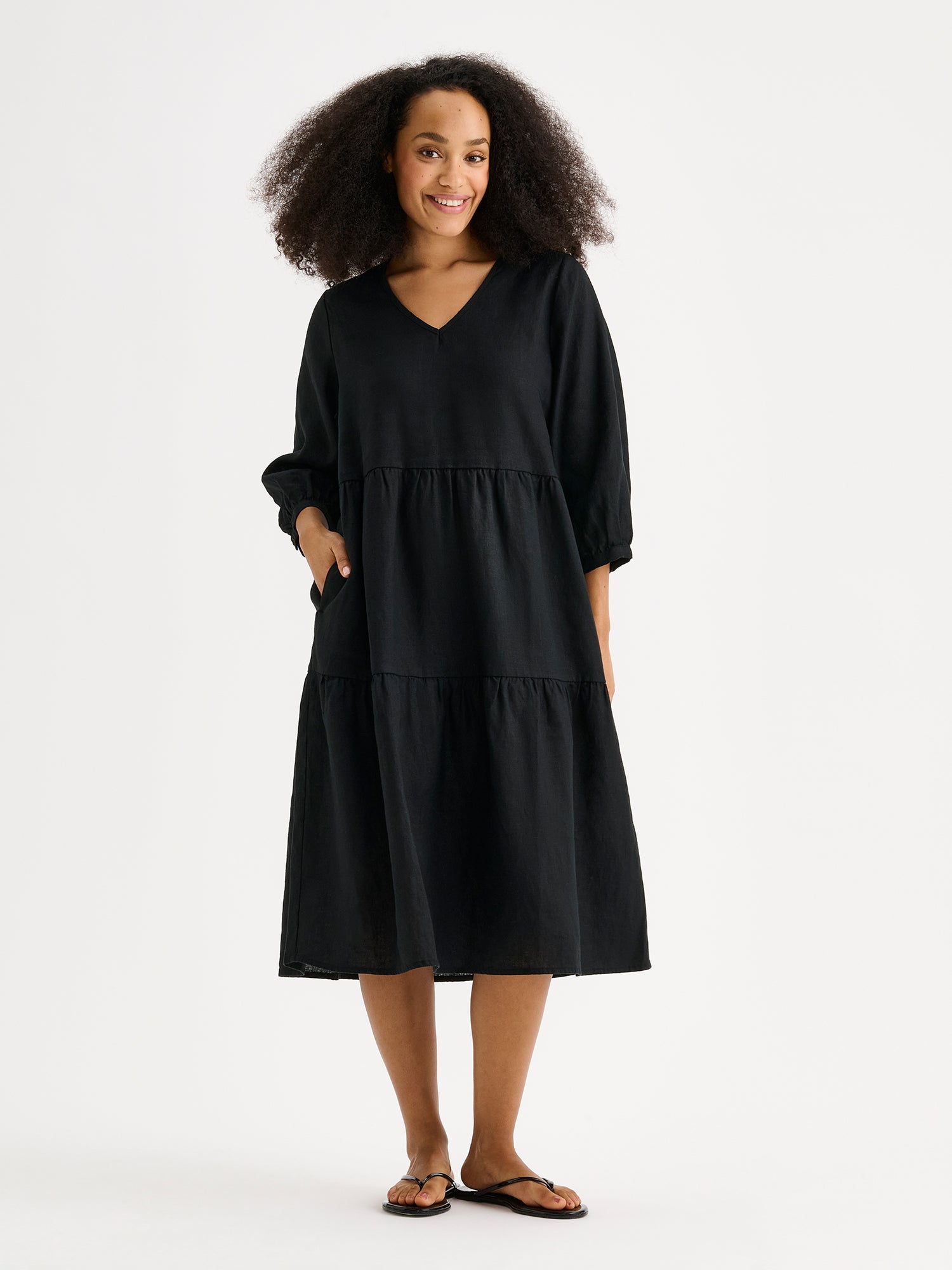 Nanso women's SARALIVIA linen dress in black features charming layered ruffles, 3/4 sleeves, side pockets and a V-neckline. Pictured on a model with the other hand in the pocket.