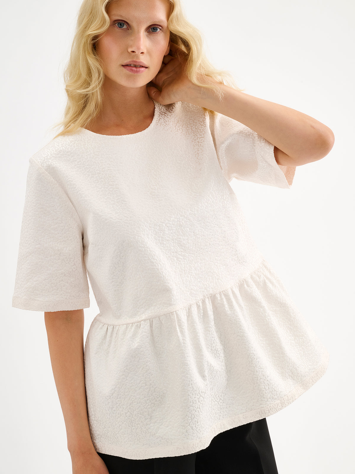 Nanso women's beautiful and elegant natural white Leora blouse captivates with its peplum-style silhouette and striking fabric, combining a subtle sheen with a richly textured surface. Pictured on a model with black trousers.