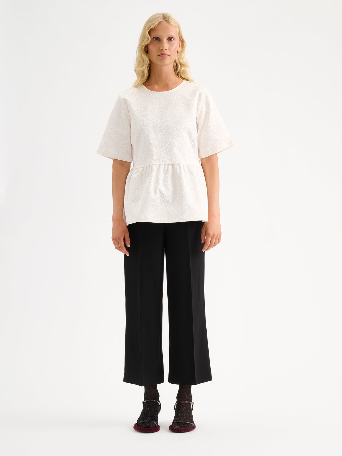Nanso women's beautiful and elegant natural white Leora blouse captivates with its peplum-style silhouette and striking fabric, combining a subtle sheen with a richly textured surface. Pictured on a model with black trousers.