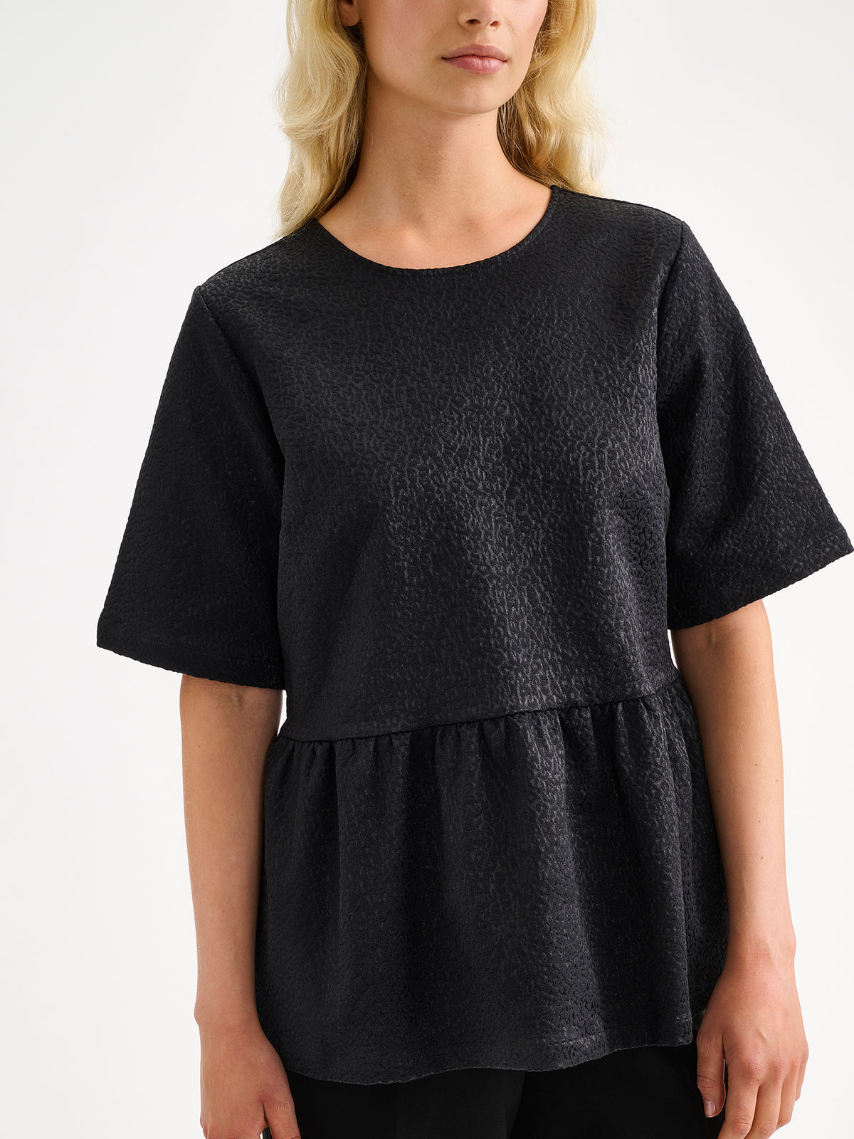 Nanso women's beautiful and elegant black Leora blouse captivates with its peplum-style silhouette and striking fabric, combining a subtle sheen with a richly textured surface, on a model with black trousers.