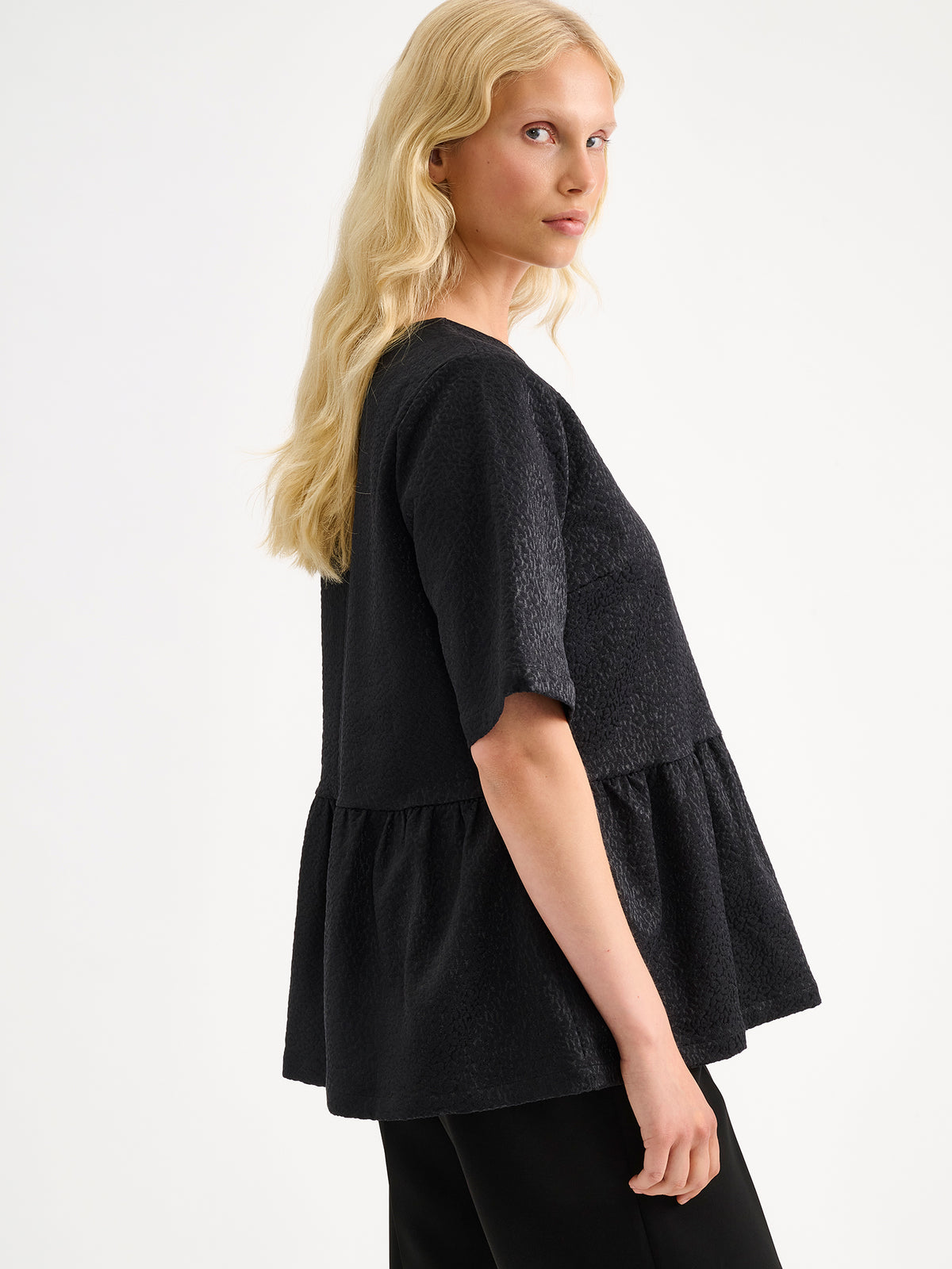 Nanso women's beautiful and elegant black Leora blouse captivates with its peplum-style silhouette and striking fabric, combining a subtle sheen with a richly textured surface, on a model with black trousers, side angle.