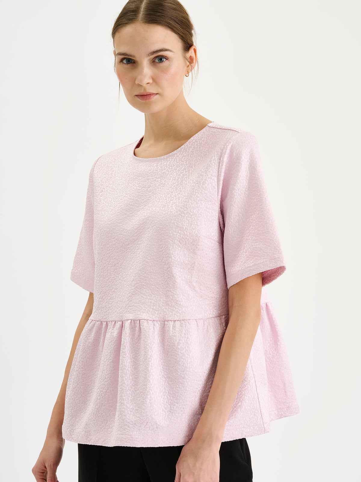 Nanso women's beautiful and elegant light pink Leora blouse captivates with its peplum-style silhouette and striking fabric, combining a subtle sheen with a richly textured surface, on a model with black trousers, close-up.