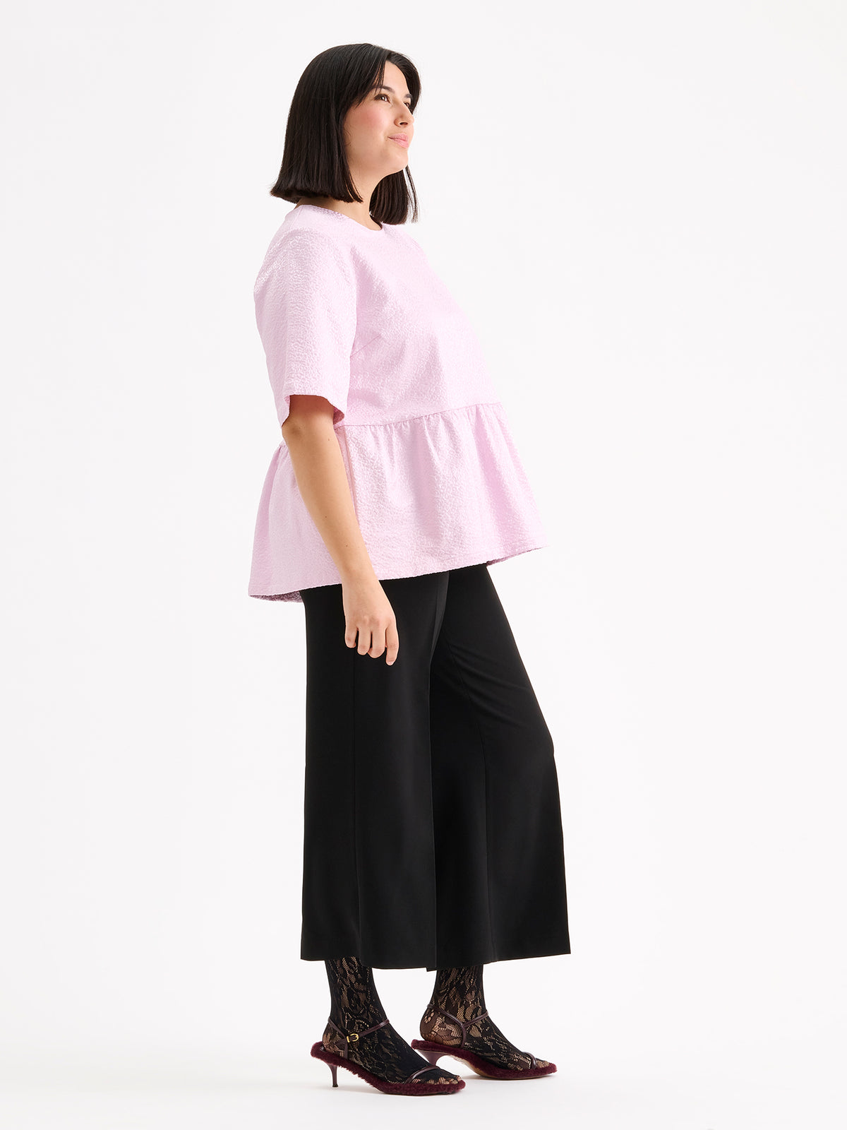 Nanso women's beautiful and elegant light pink Leora blouse captivates with its peplum-style silhouette and striking fabric, combining a subtle sheen with a richly textured surface, on a model with black trousers.