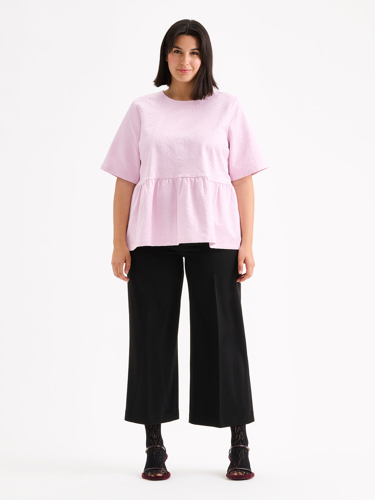 Nanso women's beautiful and elegant light pink Leora blouse captivates with its peplum-style silhouette and striking fabric, combining a subtle sheen with a richly textured surface, on a model with black trousers. | 46 - 178cm