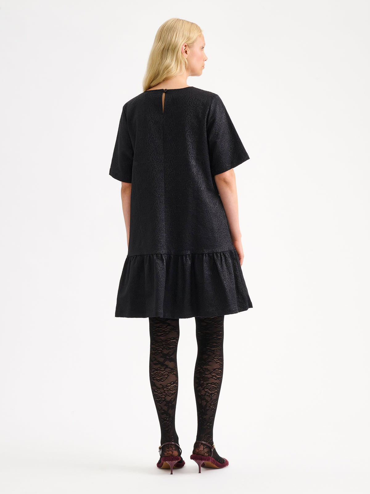 Nanso women's striking black Leora dress captivates with its beautiful fabric, combining a festive sheen with a rich textured surface, on a model with heels, pictured from the backside.