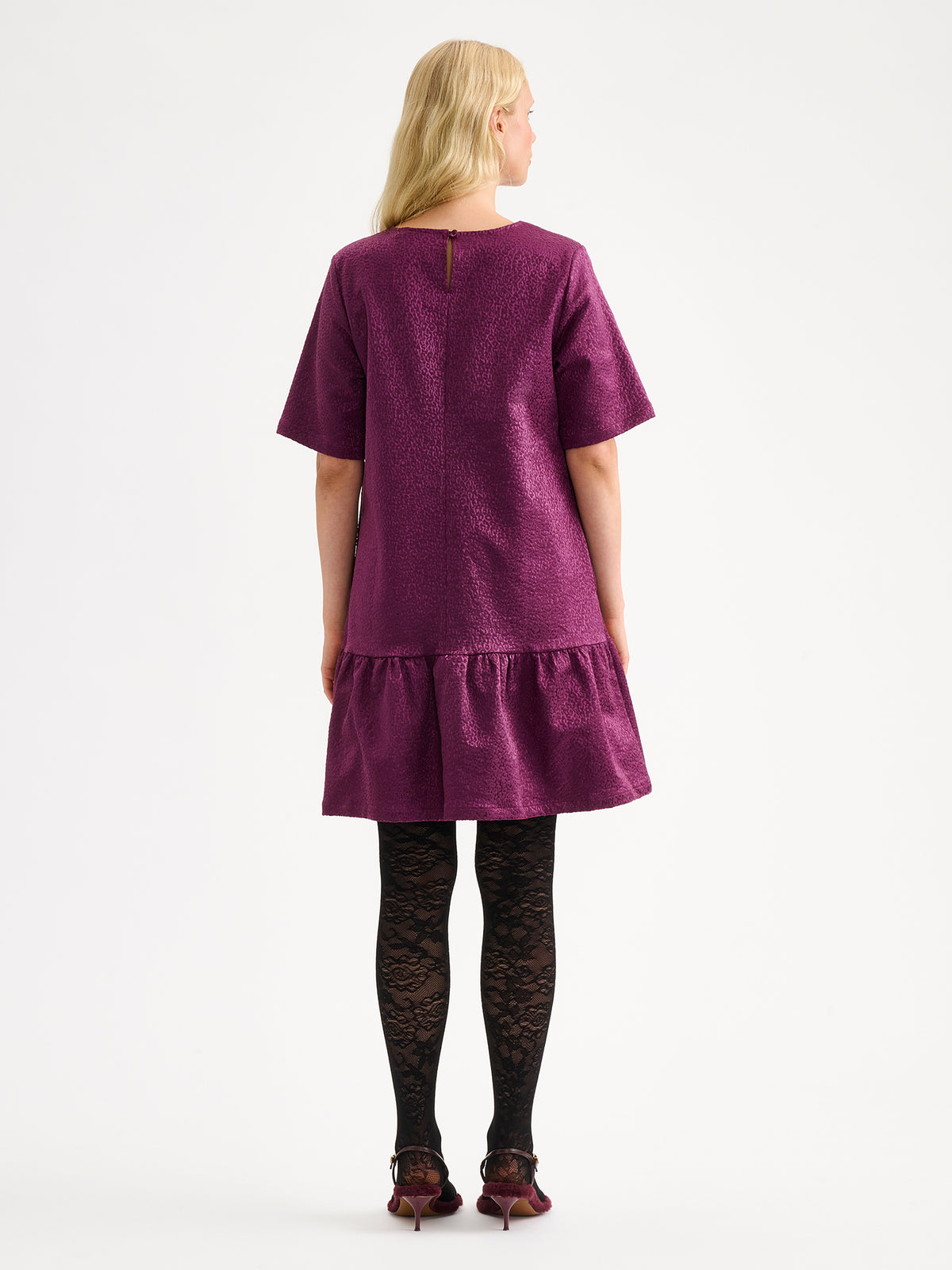 Nanso women's striking Leora dress captivates with its beautiful fabric, combining a festive sheen with a rich textured surface, pictured on a model with heels, from the backside.