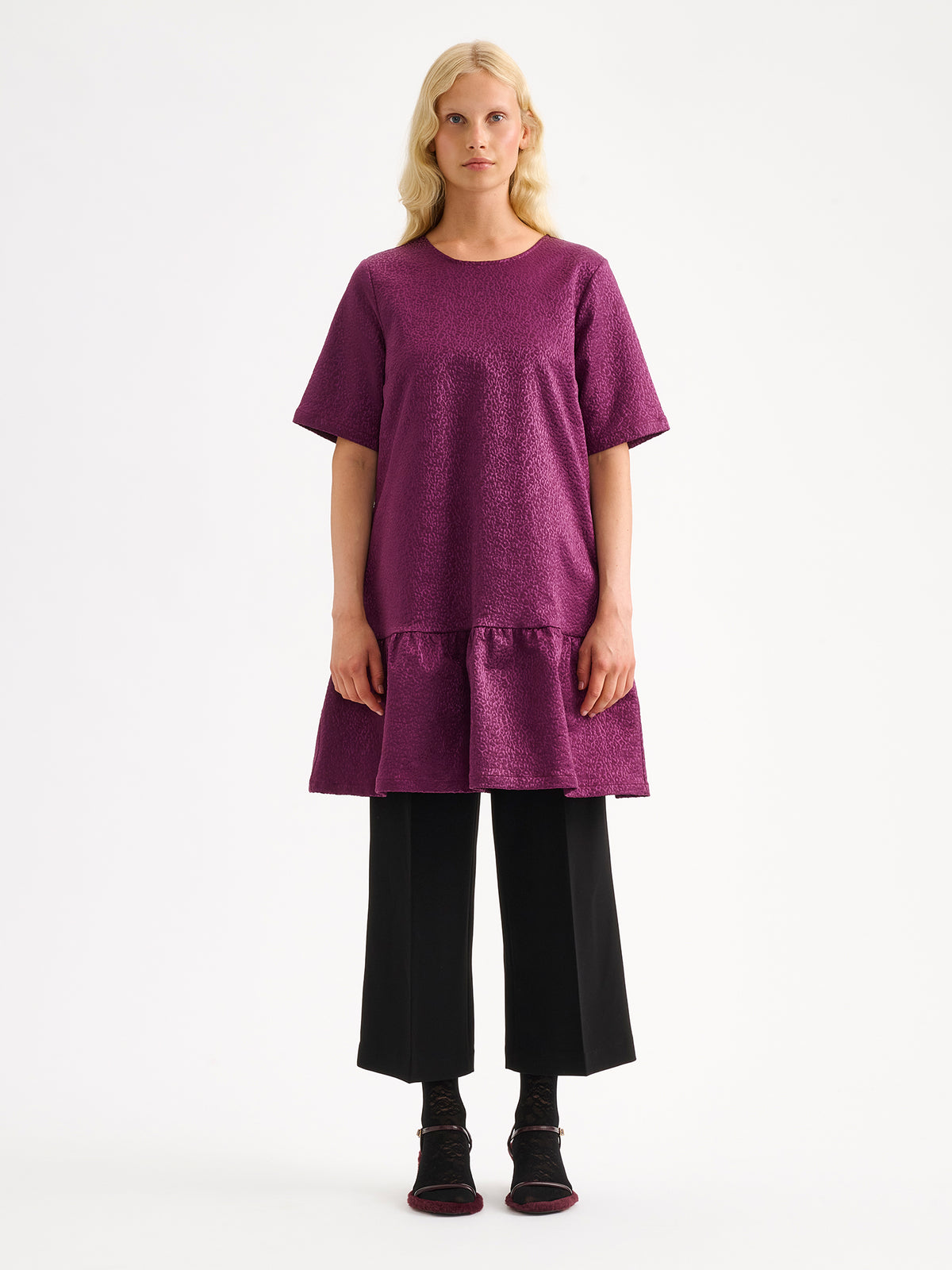 Nanso women's striking Leora dress captivates with its beautiful fabric, combining a festive sheen with a rich textured surface, pictured on a model with heels and black trousers.