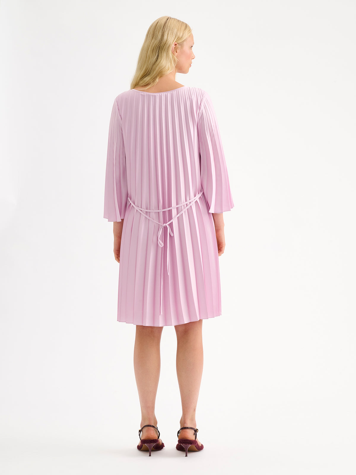 Nanso women's beautifully draping Taite dress in a deliciously cool light pink shade. This versatile pleated dress can be worn with or without the slim belt. This A-line dress features 3/4-length sleeves and a slim belt at the waist. Pictured on a model with heels, from the backside.