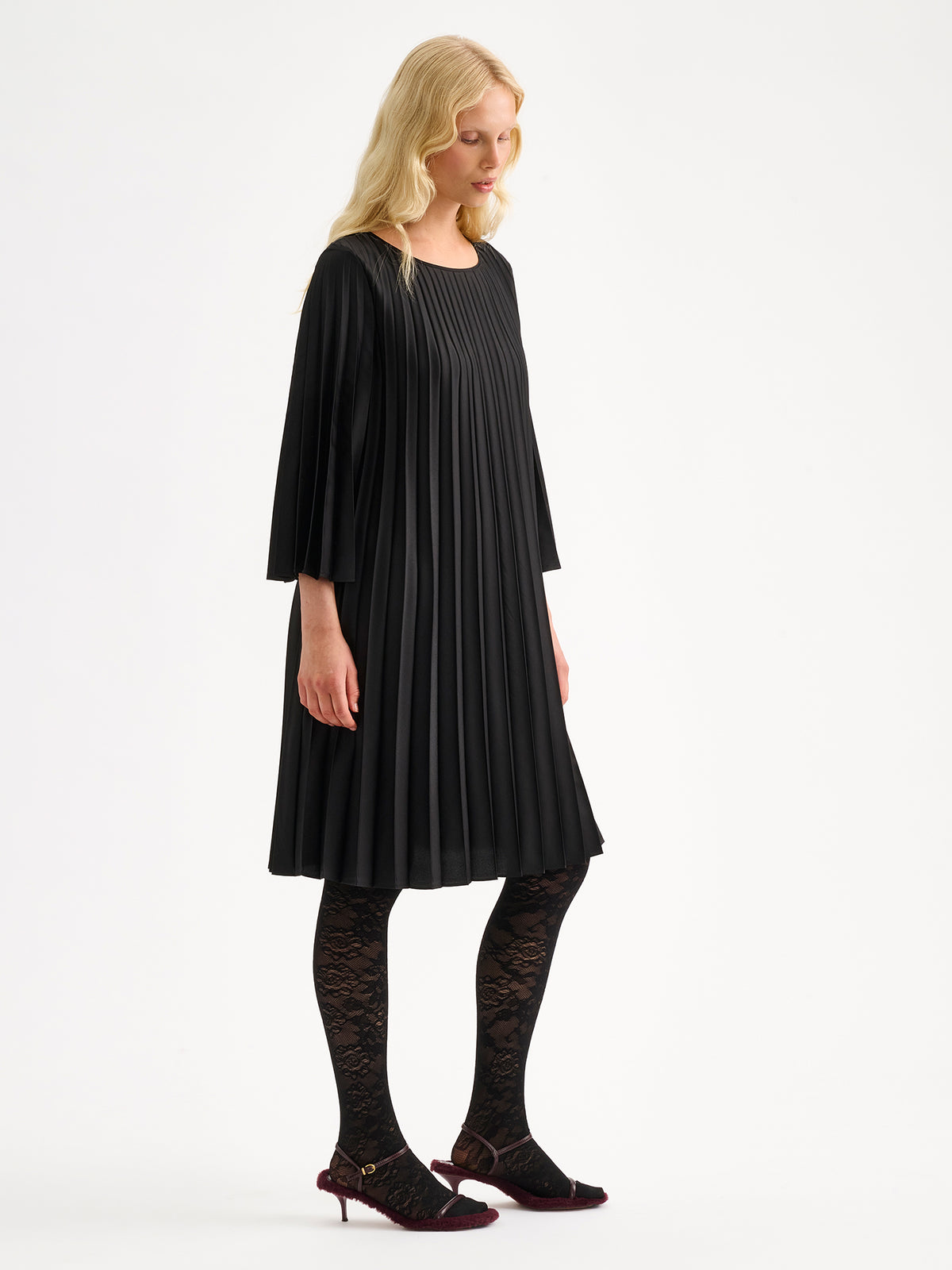 Nanso women's beautifully draping Taite dress in a classic black shade. This versatile pleated dress can be worn with or without the slim belt. This A-line dress features 3/4-length sleeves and a slim belt at the waist. Pictured on a model with black lace tights without the belt and from the side.