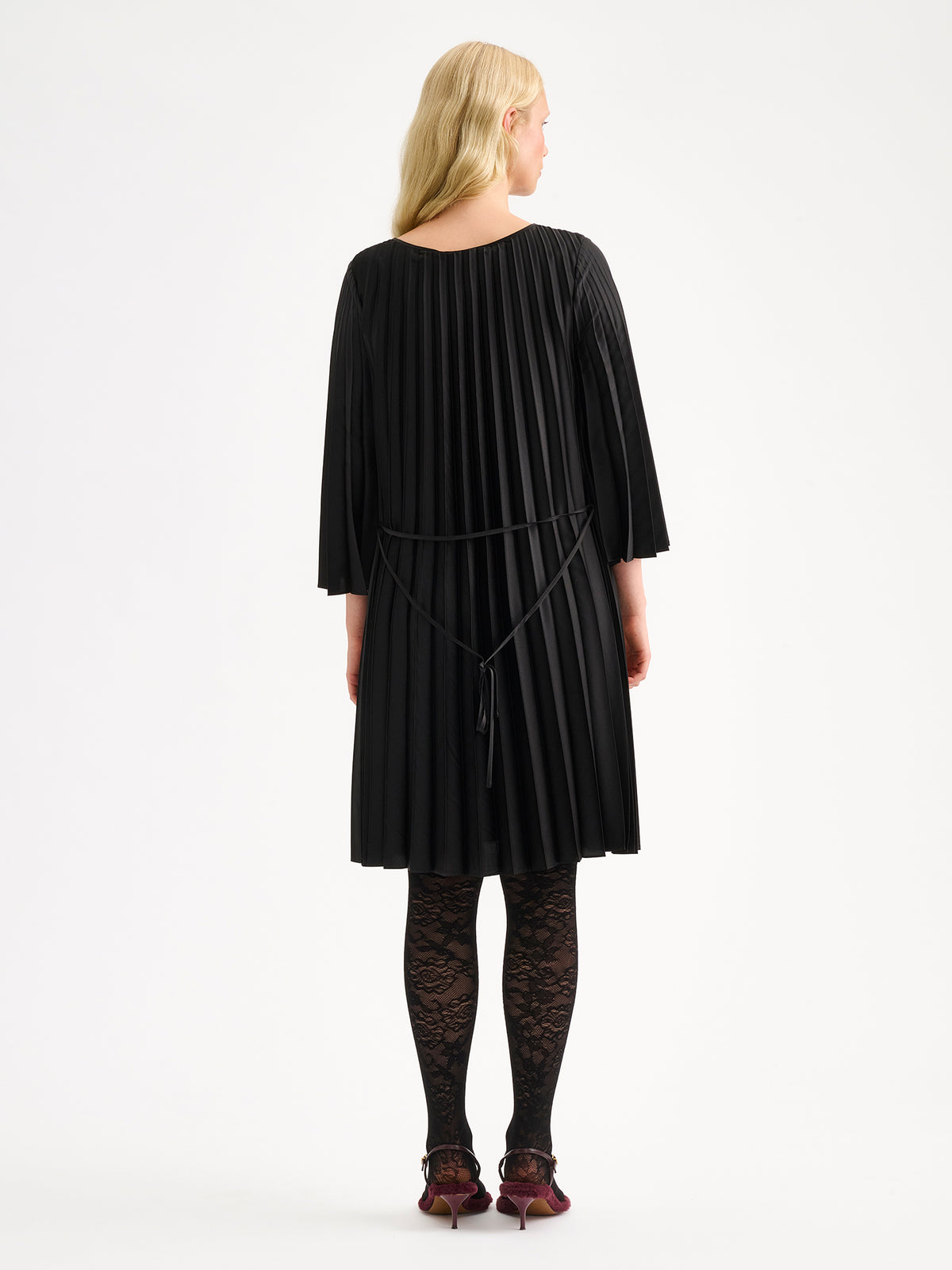 Nanso women's beautifully draping Taite dress in a classic black shade. This versatile pleated dress can be worn with or without the slim belt. This A-line dress features 3/4-length sleeves and a slim belt at the waist. Pictured on a model with black lace tights, from the backside.