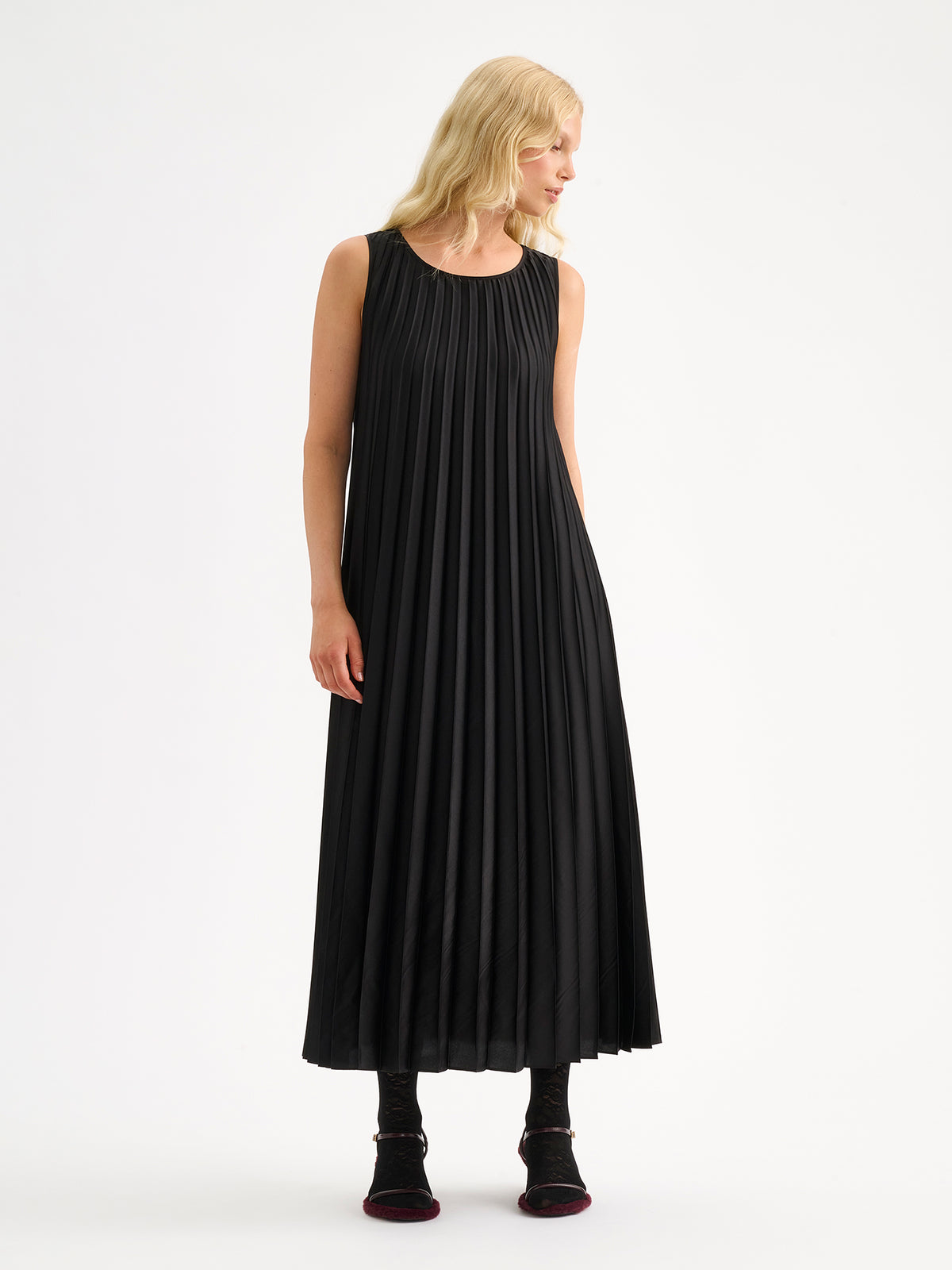 Nanso women's beautifully draping long Taite dress in a classic black shade. This long, sleeveless pleated dress with a slim belt is a striking choice for festive occasions and other special moments throughout the season. Pictured on a model with heels without the belt.