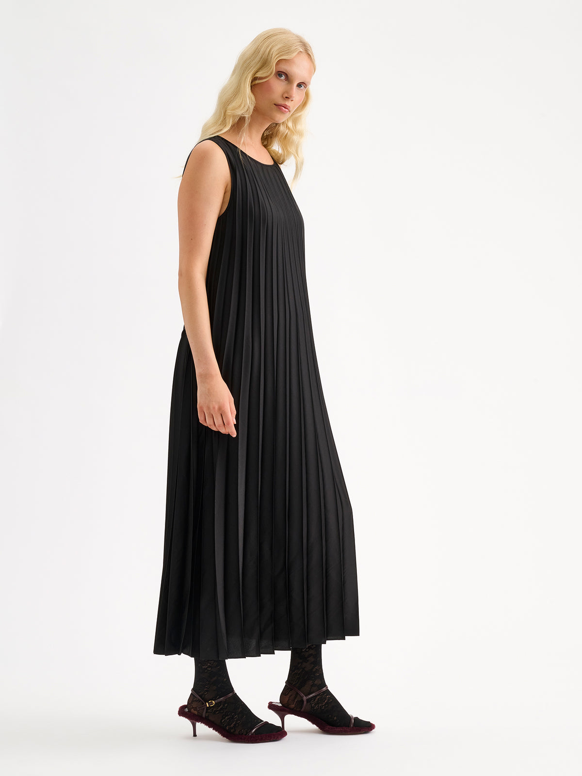Nanso women's beautifully draping long Taite dress in a classic black shade. This long, sleeveless pleated dress with a slim belt is a striking choice for festive occasions and other special moments throughout the season. Pictured on a model with heels, from the side angle.