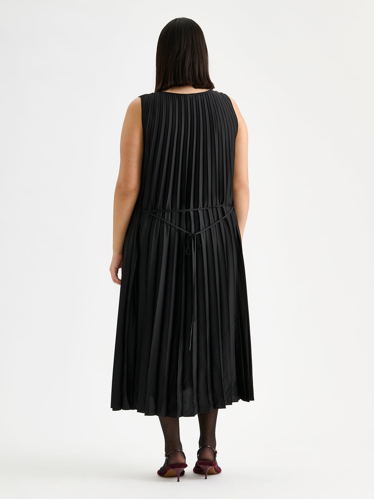 Nanso women's beautifully draping long Taite dress in a classic black shade. This long, sleeveless pleated dress with a slim belt is a striking choice for festive occasions and other special moments throughout the season. Pictured on a model with heels, from the backside.