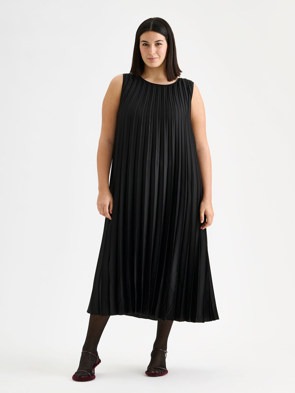 Nanso women's beautifully draping long Taite dress in a classic black shade. This long, sleeveless pleated dress with a slim belt is a striking choice for festive occasions and other special moments throughout the season. Pictured on a model with heels.