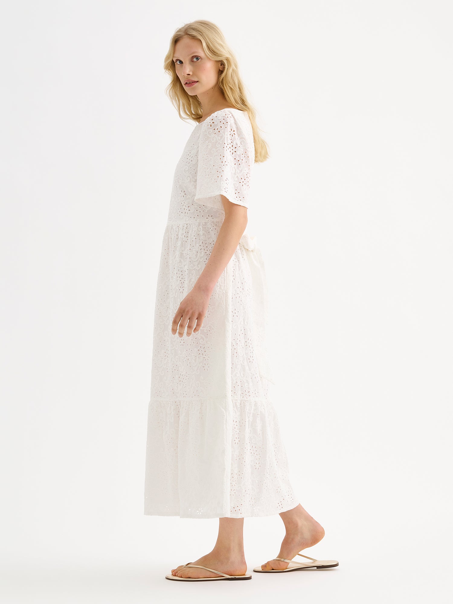 Nanso women's beautiful long soft white Odette dress crafted from delicate broderie anglaise style cotton fabric with a lace-like texture on a model with sandals, pictured from the side.