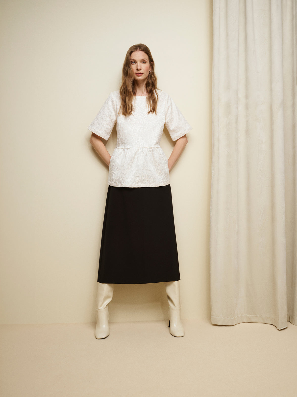 Nanso women's beautiful and elegant natural white Leora blouse captivates with its peplum-style silhouette and striking fabric, combining a subtle sheen with a richly textured surface. Pictured on a model with a black midi skirt. | 36 - 175,5cm