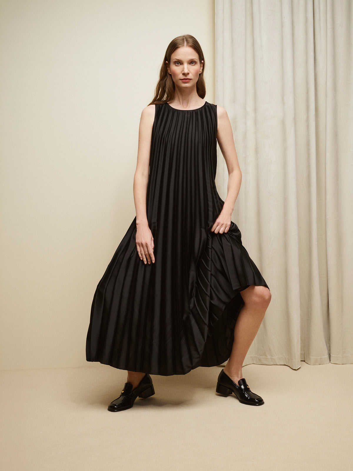 Nanso women's beautifully draping long Taite dress in a classic black shade. This long, sleeveless pleated dress with a slim belt is a striking choice for festive occasions and other special moments throughout the season. Pictured on a model with black shoes.
