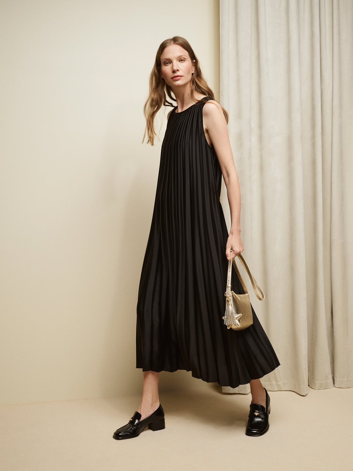 Nanso women's beautifully draping long Taite dress in a classic black shade. This long, sleeveless pleated dress with a slim belt is a striking choice for festive occasions and other special moments throughout the season. Pictured on a model with black shoes and bag in gold shade.