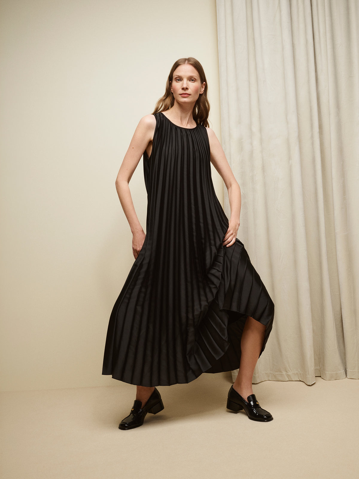 Nanso women's beautifully draping long Taite dress in a classic black shade. This long, sleeveless pleated dress with a slim belt is a striking choice for festive occasions and other special moments throughout the season. Pictured on a model with black shoes.