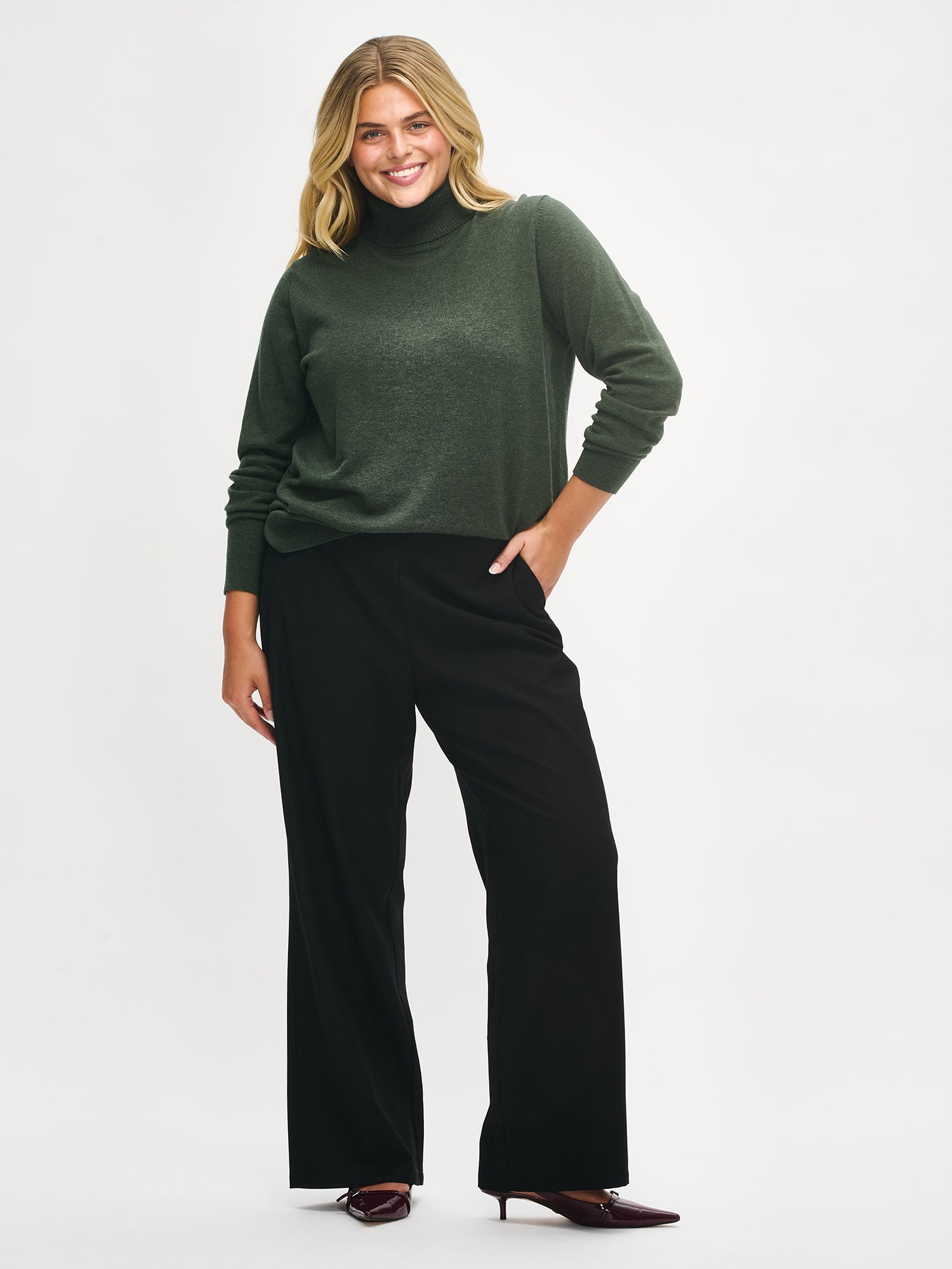 Nanso women’s dark green Villis turtleneck sweater in a merino wool and cotton blend, lightweight and timeless, full-length sleeves and hip-length hem, worn by model in full-body view.