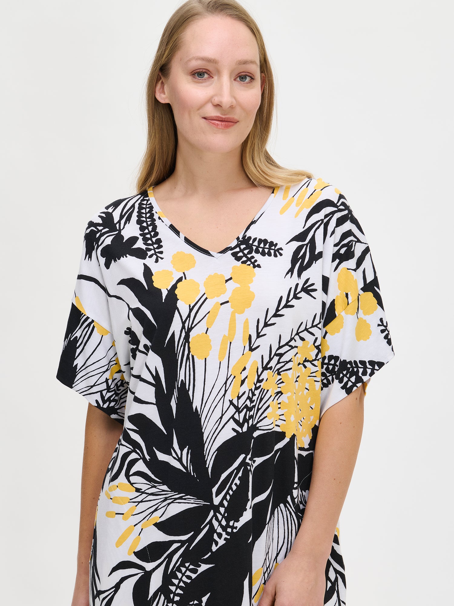 Nanso women's long floral-patterned Puutarha kaftan with black and yellow flowers on a white background, featuring a V-neckline, side pockets, and side slits, worn by the model in a close-up front view.
