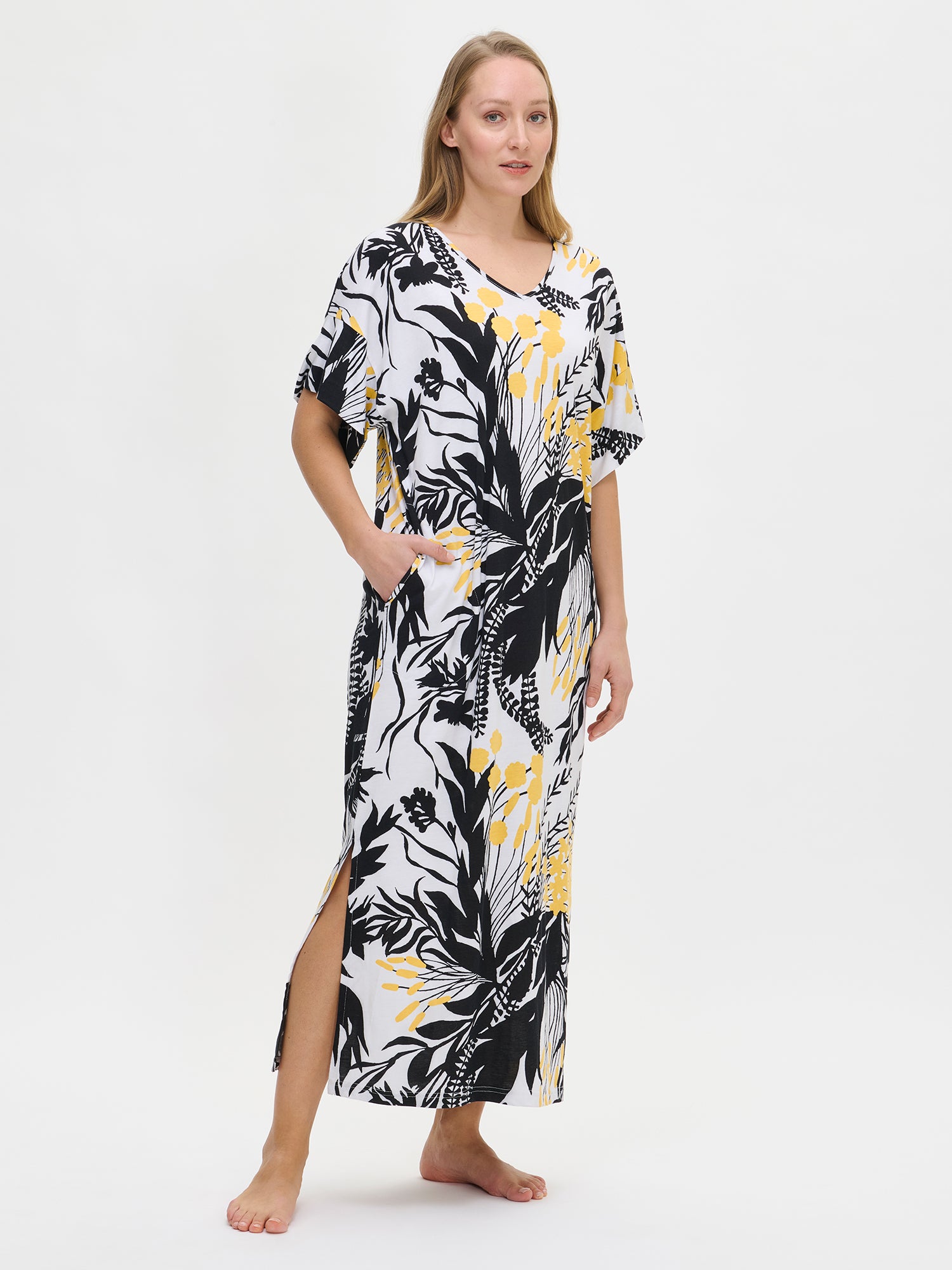 Nanso women's long floral-patterned Puutarha kaftan with black and yellow flowers on a white background, featuring a V-neckline, side pockets, and side slits, worn by the model.