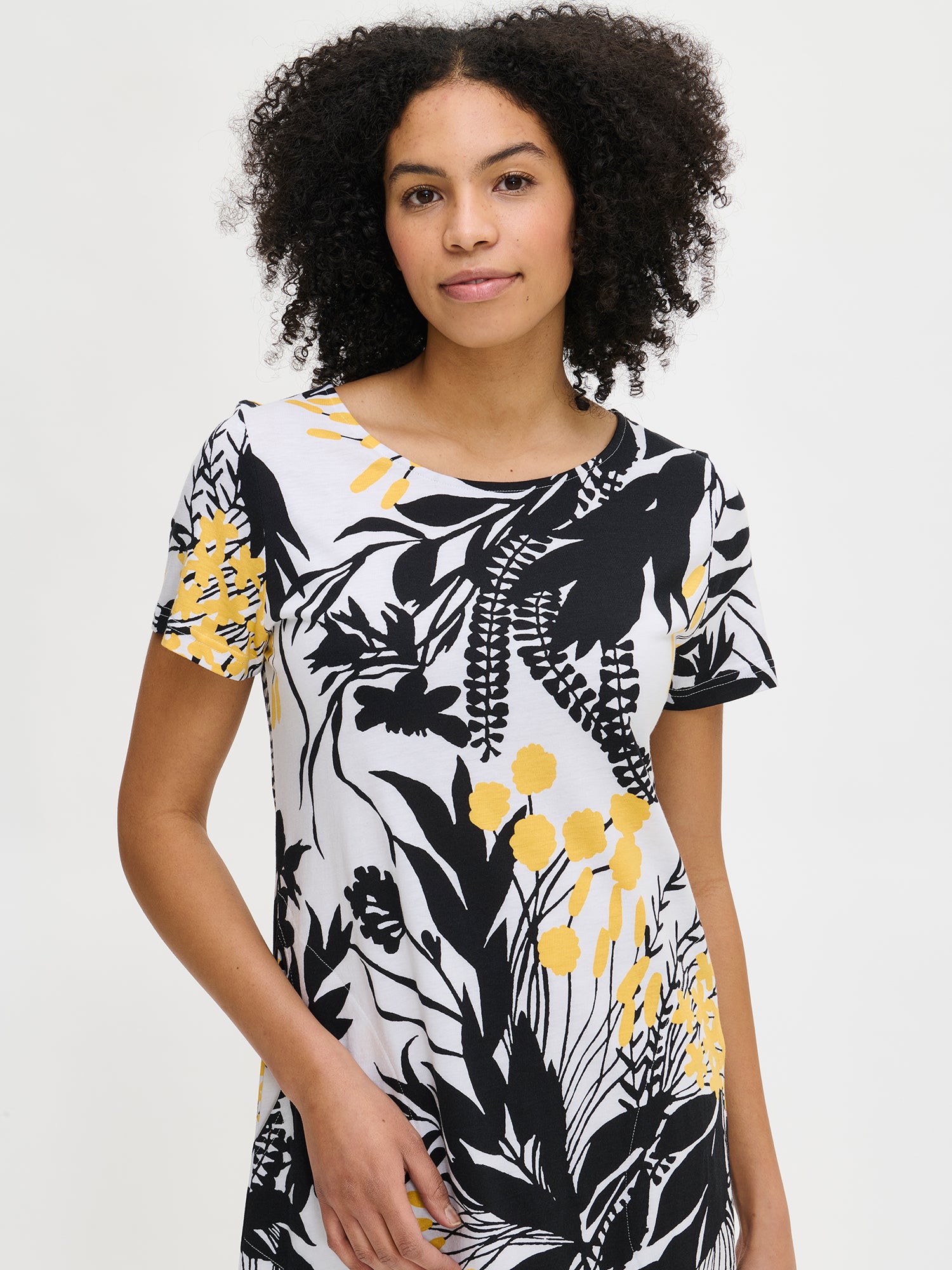Nanso women’s floral-patterned knee-length Puutarha nightgown with a white background featuring black and yellow leaves and flowers. The nightgown has short sleeves, side pockets, and an oval neckline, worn by the model in a close-up front view.