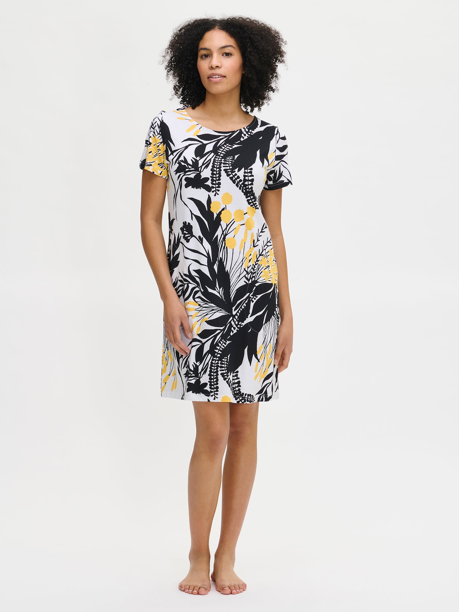Nanso women’s floral-patterned knee-length Puutarha nightgown with a white background featuring black and yellow leaves and flowers. The nightgown has short sleeves, side pockets, and an oval neckline, worn by the model.