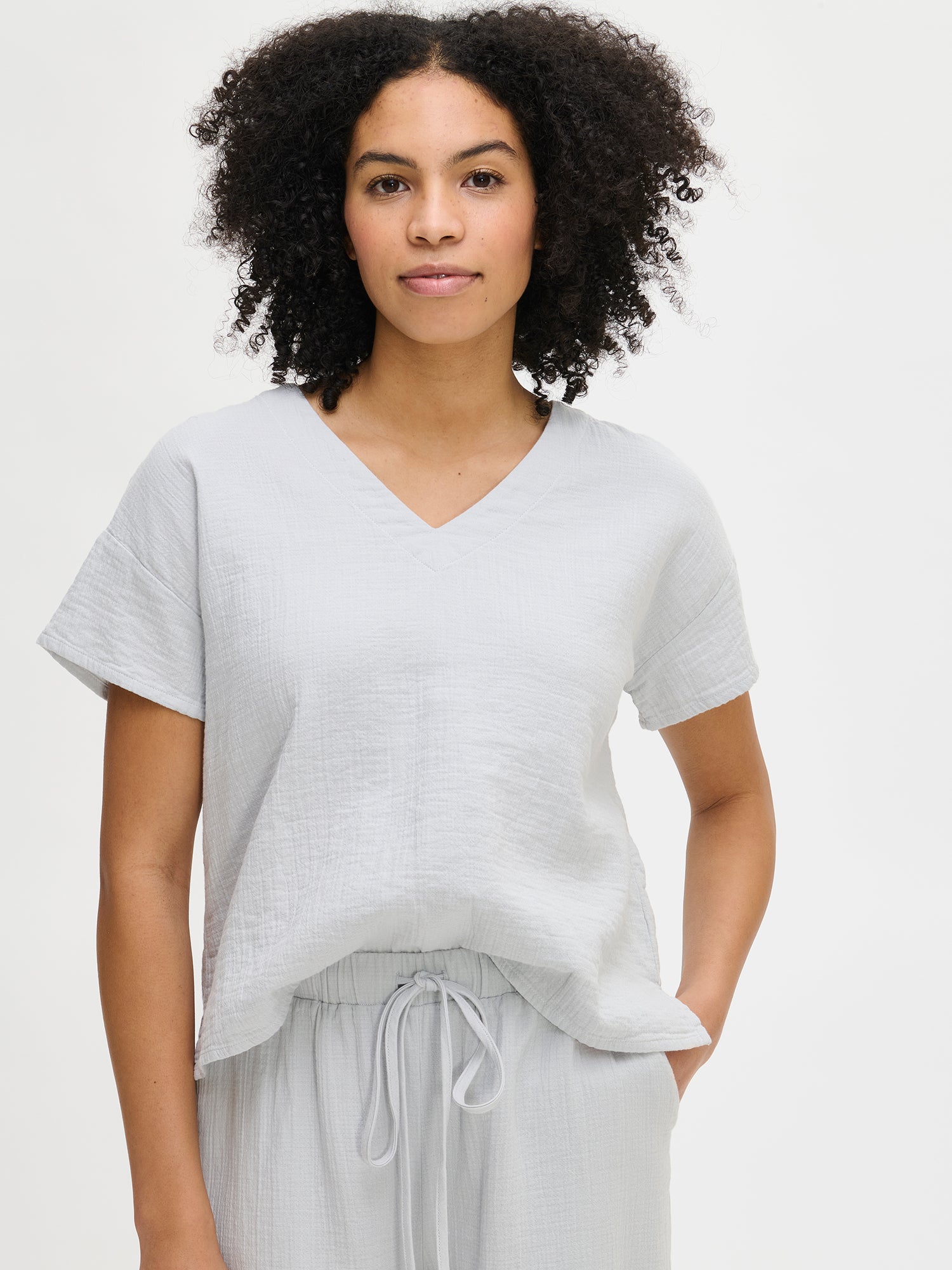Nanso women's relaxed solid blue boxy waist-length Aamu top with short sleeves, a V-neckline, a straight hem, and side slits, worn by the model in a close-up front view.