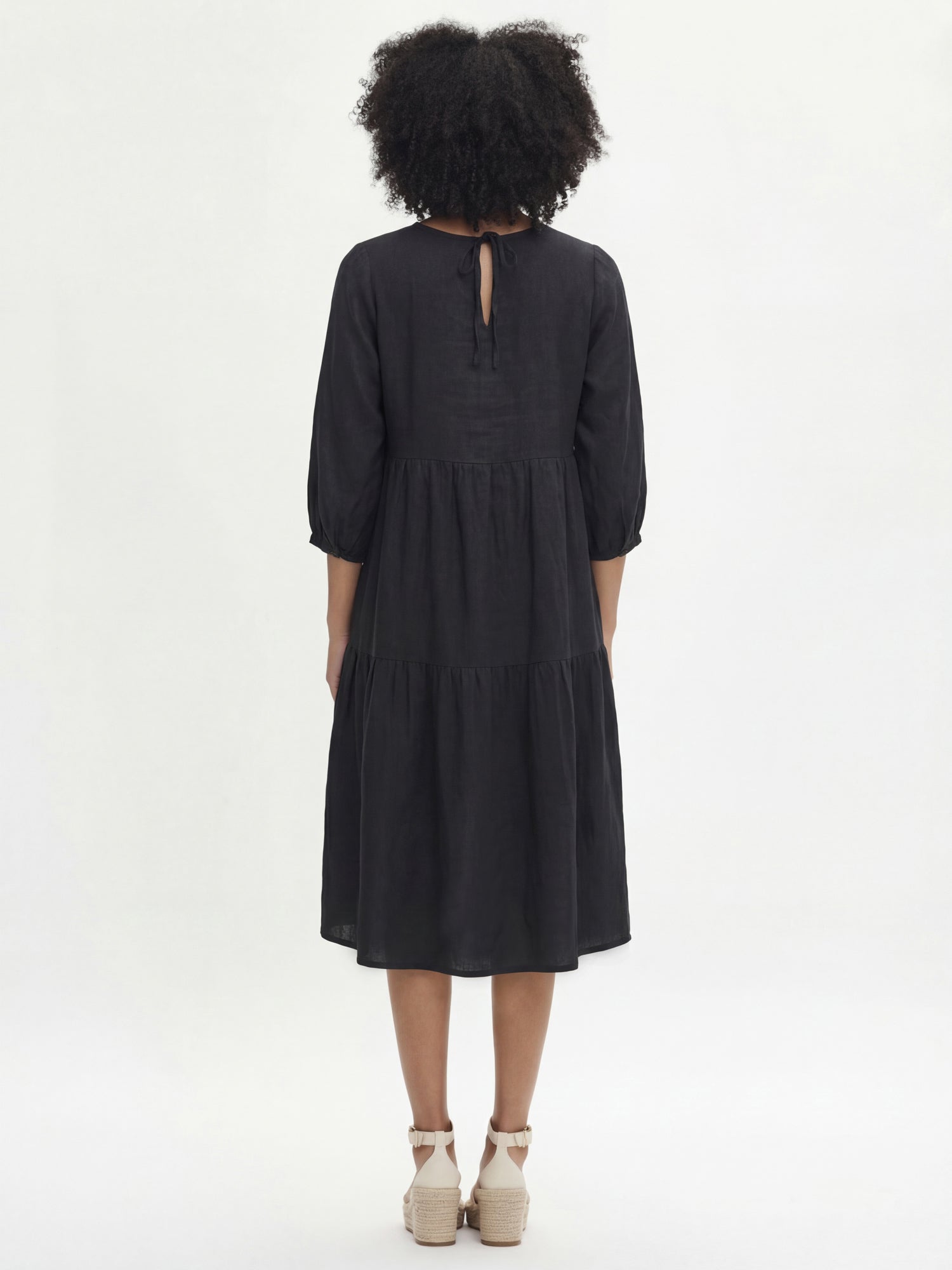 Women's Saralia Linen Dress in black features voluminous frill layers, 3/4 length sleeves that gracefully gather above the cuff with button adjusters for a tailored fit. Pictured on a model, from the backside