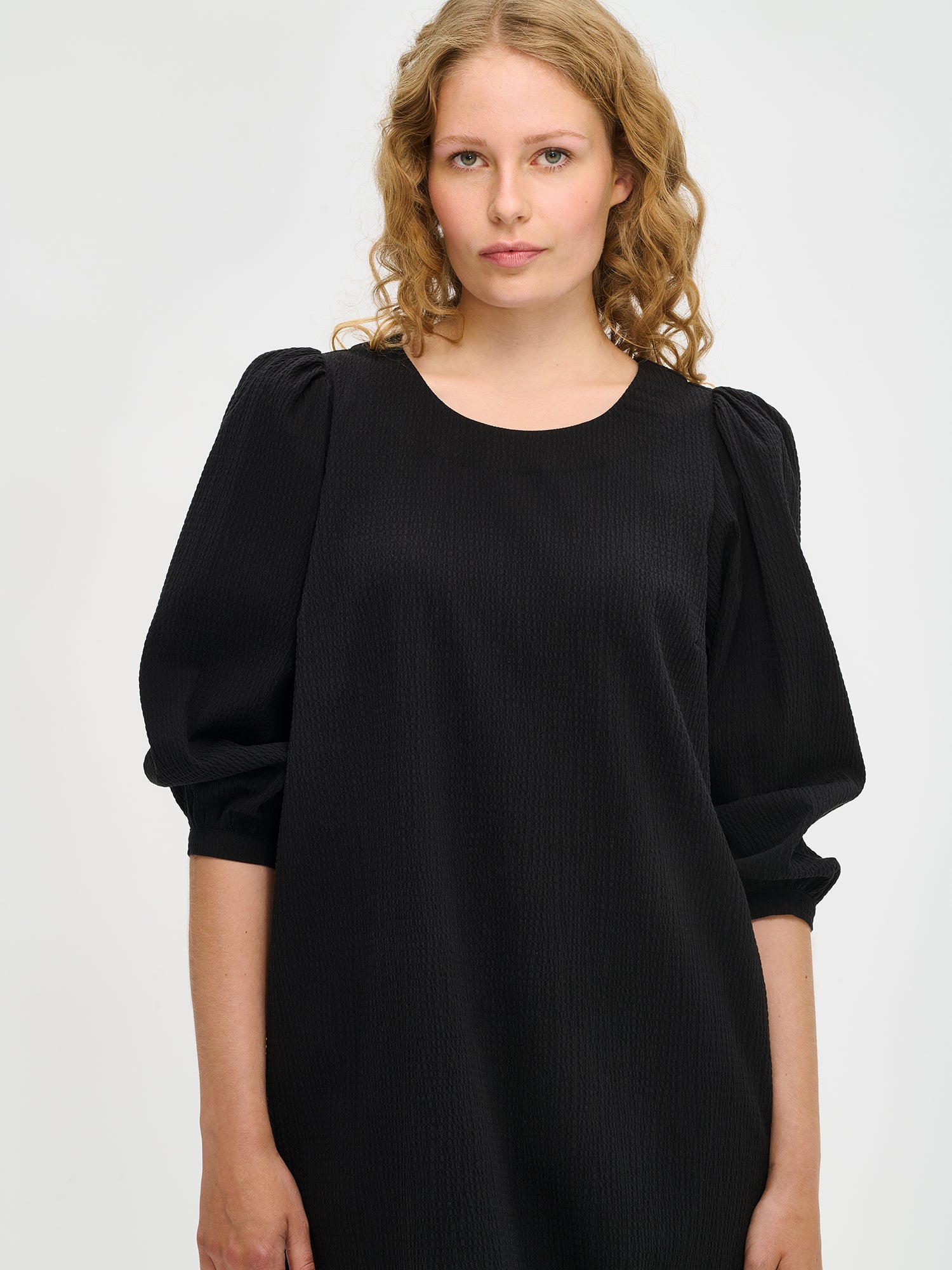 Nanso women’s solid black Bianca dress in cotton–elastane, round neckline and gathered sleeves, knee-length, worn by model in close-up view.