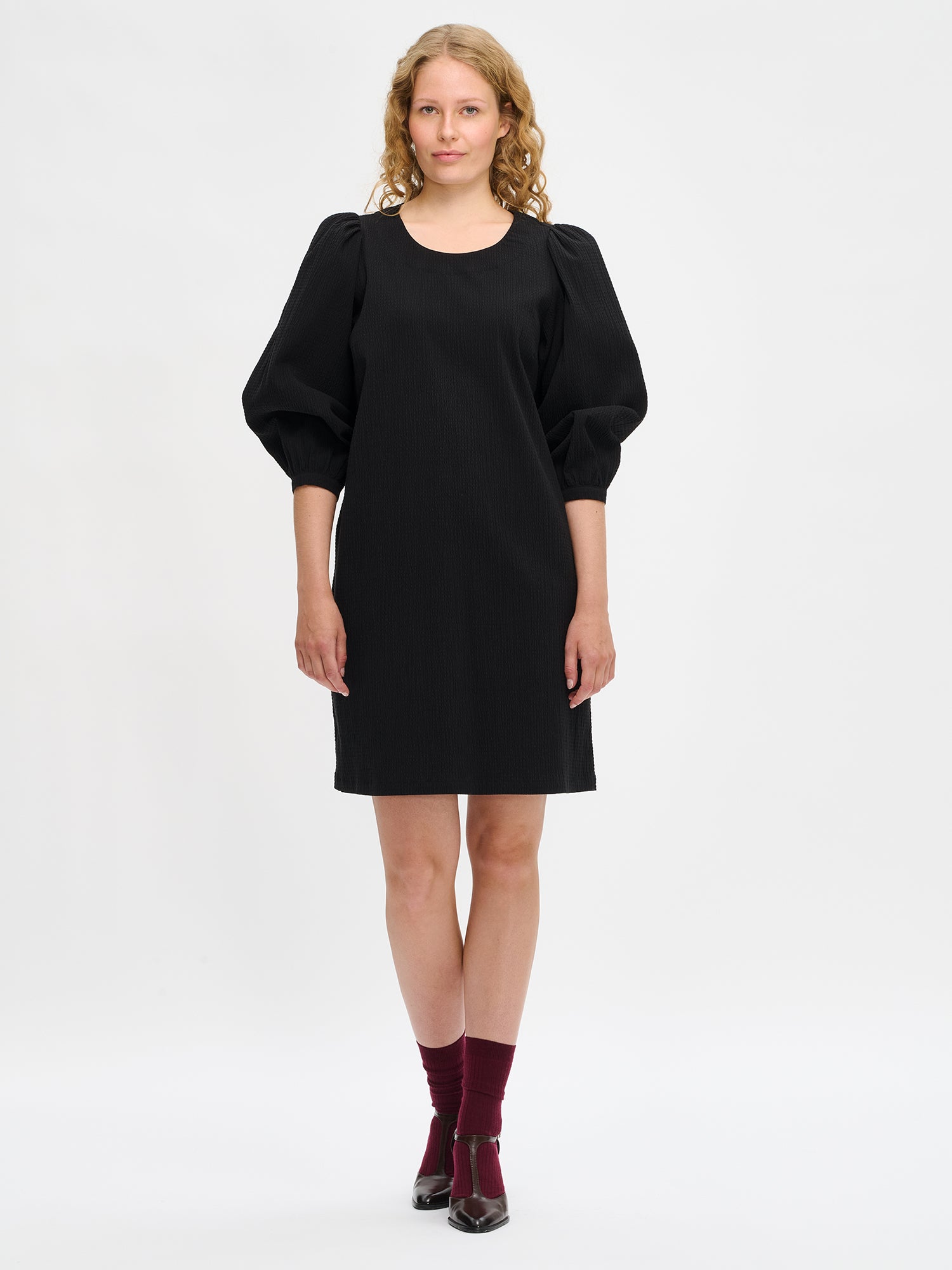 Nanso women’s solid black Bianca dress in cotton–elastane, round neckline, gathered 3/4 sleeves and pockets, knee-length, worn by model in full-body view.