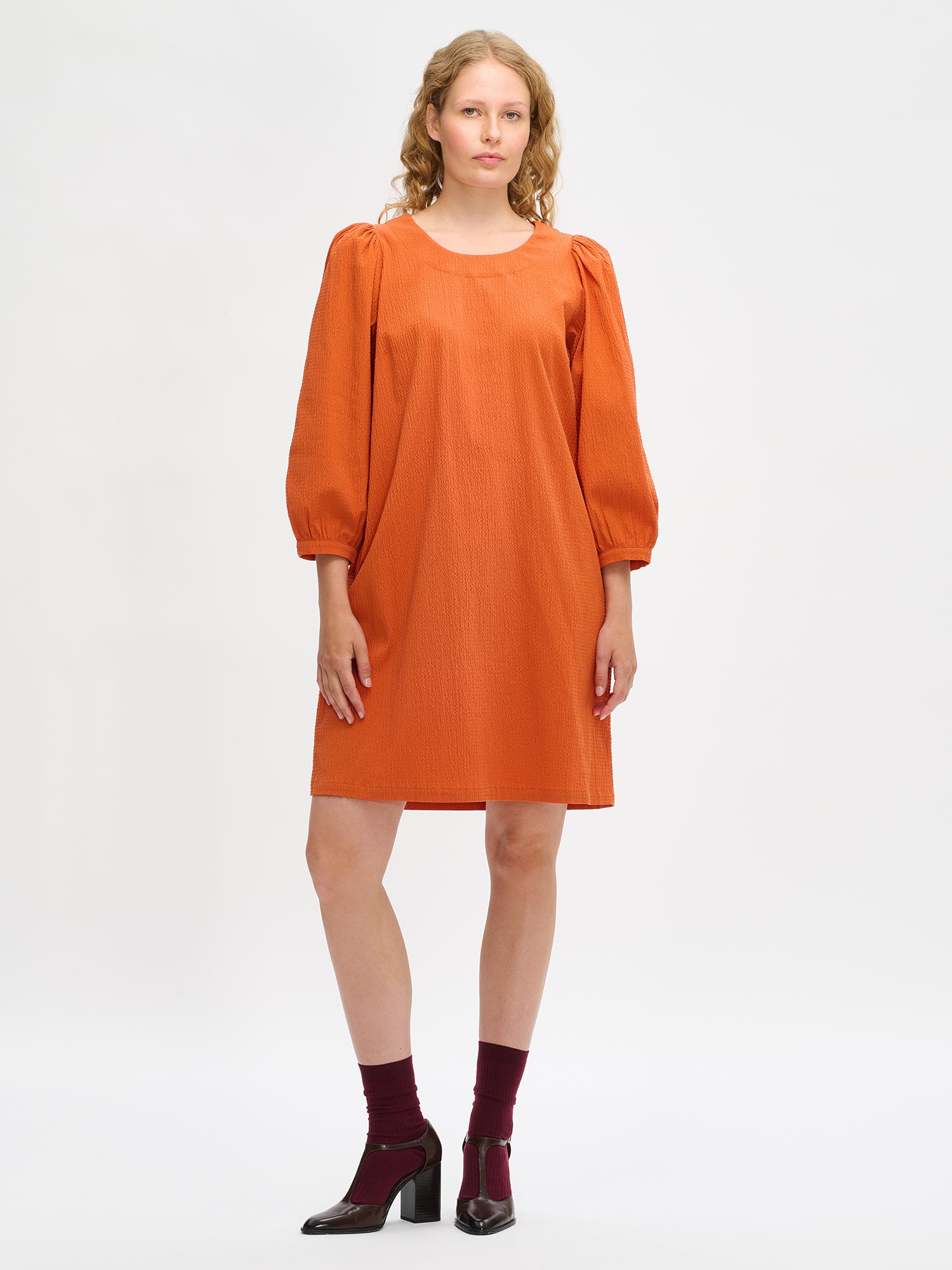 Nanso women’s solid orange Bianca dress in cotton–elastane, round neckline, gathered 3/4 sleeves and pockets, knee-length, worn by model in full-body view.