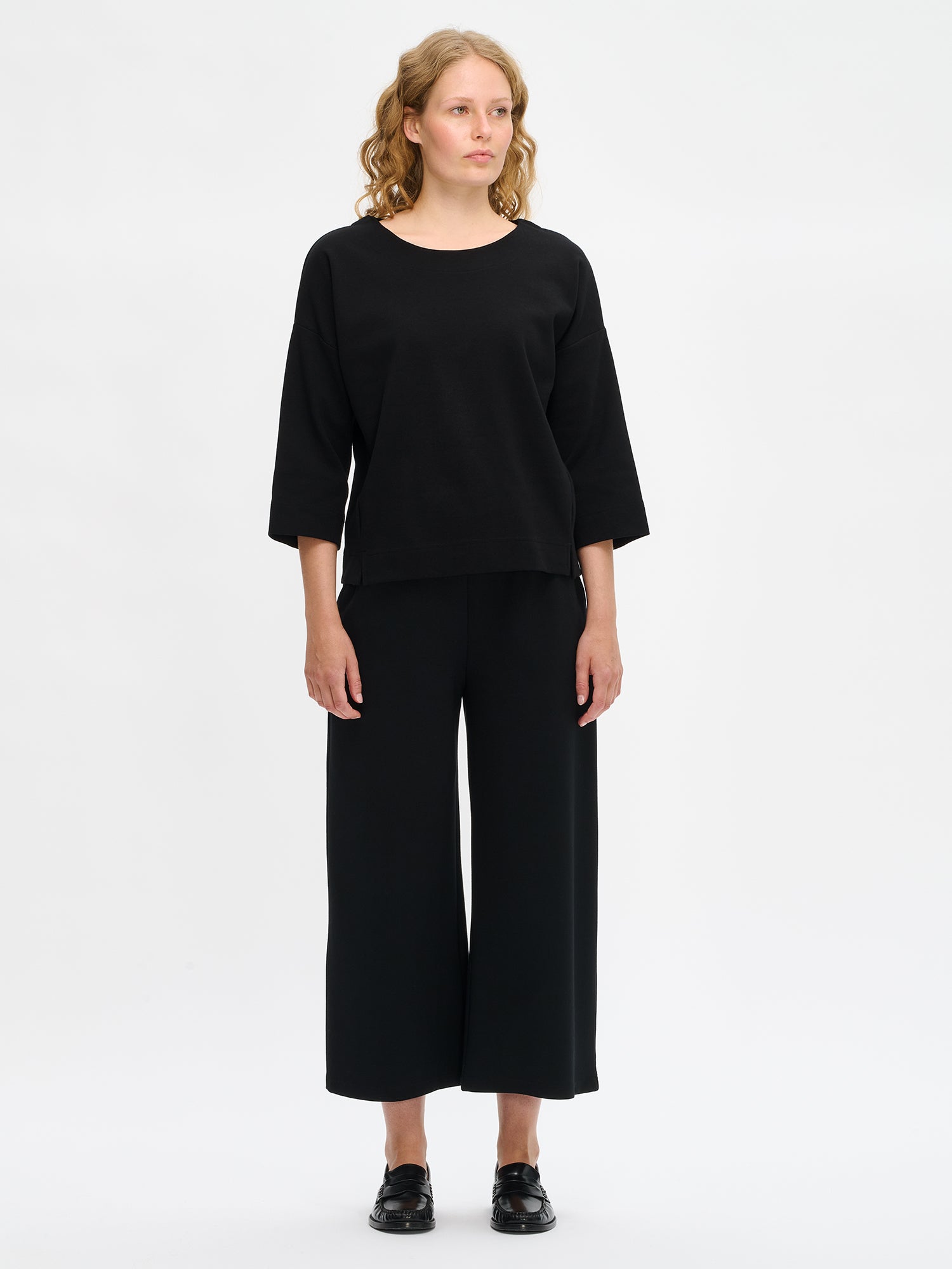 Nanso women’s black Elena top in cotton–polyester–elastane knit, oval neckline, 3/4 sleeves and dropped shoulders, worn by model in full-body view.