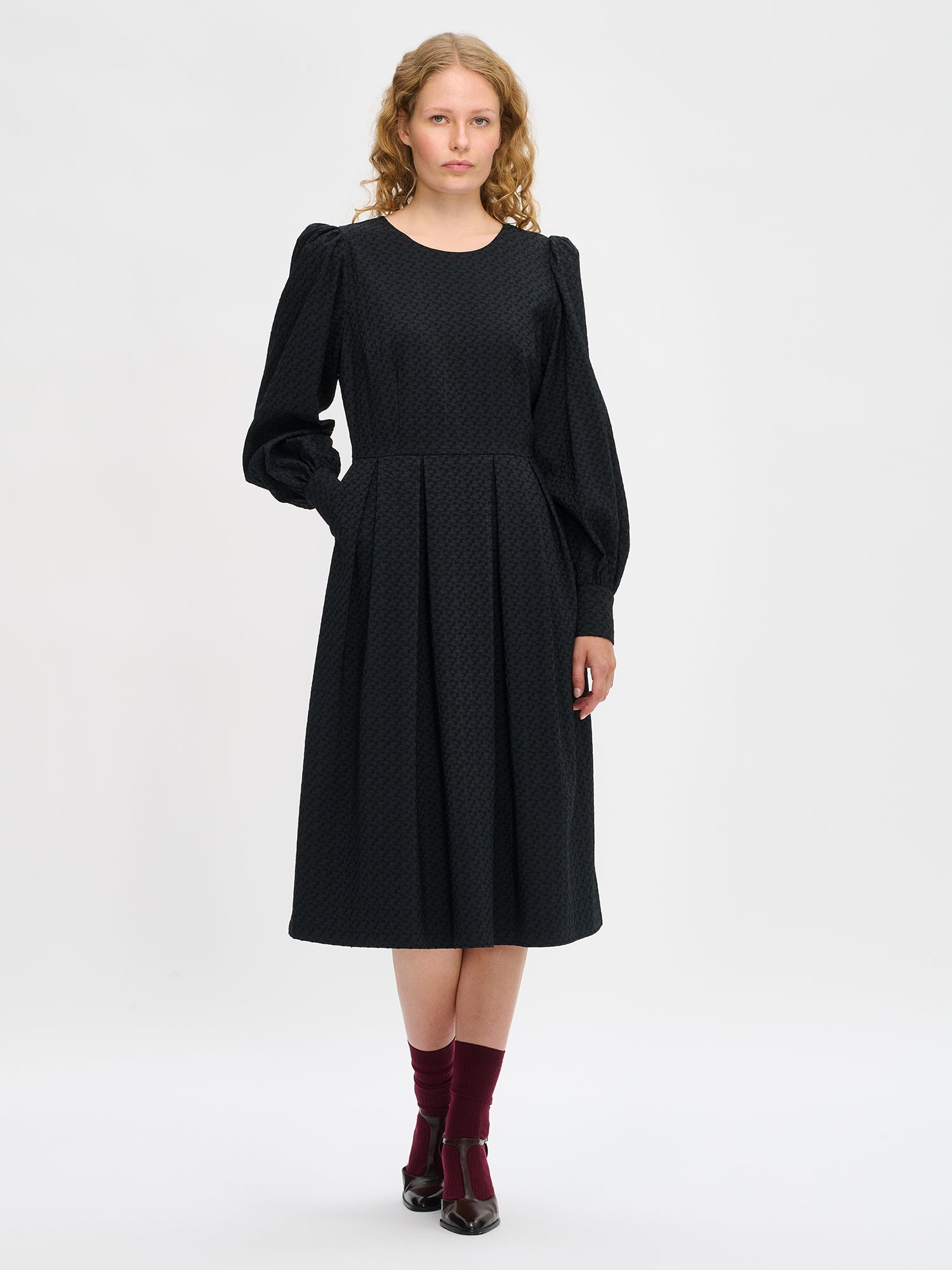 Nanso women’s black Reliefi dress, midi-length style with round neckline, long gathered sleeves and pleated waist, worn by model in full-body photo.