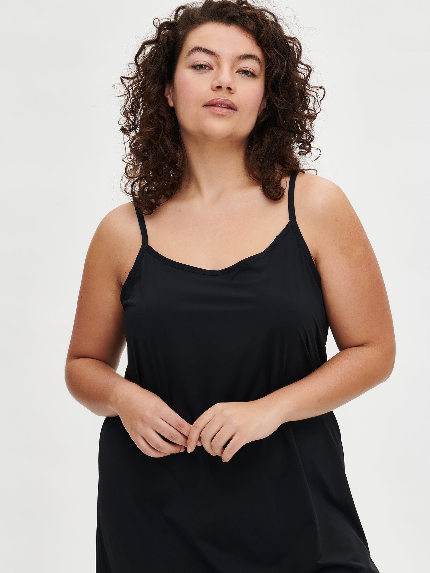 Nanso women’s black Basic slip dress with spaghetti straps, mid-length front view on the plus size model. Hands held together.