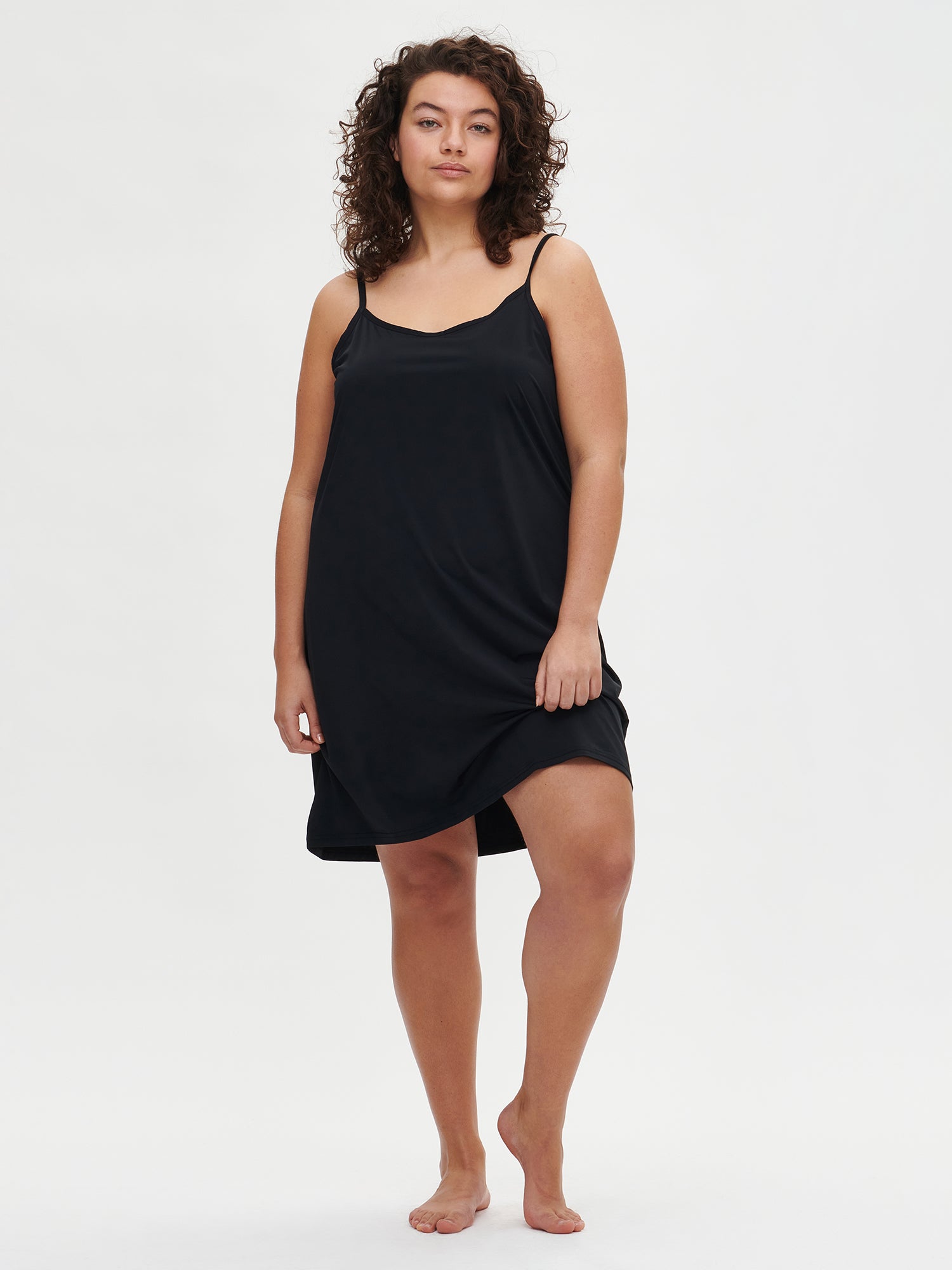 Nanso women’s black Basic slip dress with spaghetti straps, front view on the plus size model. The hem is lifted slightly to show movement.