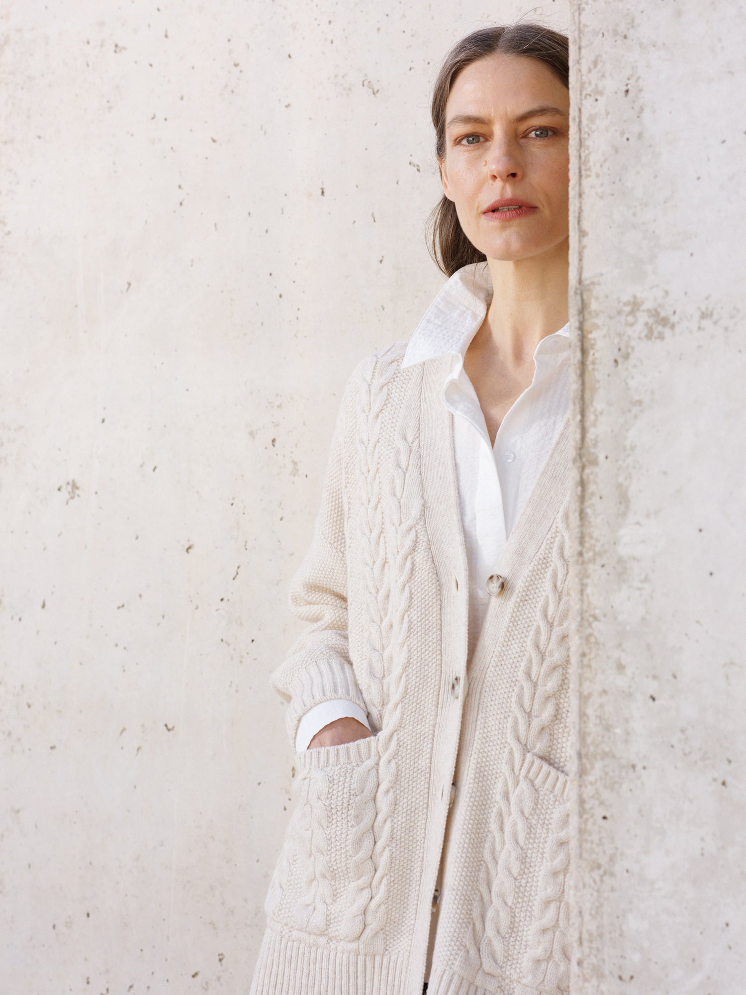 Woman wearing a cream cable knit cardigan over a white shirt against a light stone wall.