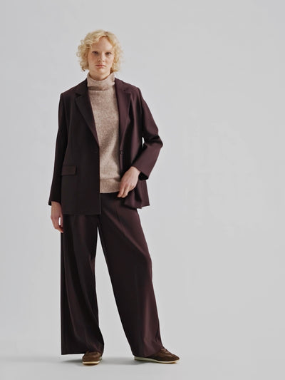 A stylish women's Eeva blazer in a rich brown shade with a relaxed yet structured silhouette, on a model with a beige knitted vest and sneakers | 36 - 177cm